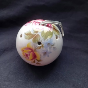 Könnte beinhalten: Weißer, kugelförmiger Keramikschmuck mit einem Blumendesign mit rosa, gelben und blauen Rosen. Der Schmuck hat drei kleine Löcher und eine silberfarbene Metallschlaufe zum Aufhängen. Der Hintergrund ist ein dunkelblauer Stoff.
