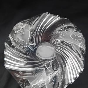 Könnte beinhalten: Dekorative Schüssel aus klarem Glas mit einem Wirbeldesign. Die Schüssel zeigt geätzte Bilder von Hirschen und floralen Mustern. Die Schüssel hat eine kreisförmige Form mit einer leicht erhöhten Mitte und einem wellenförmigen Rand. Die Schüssel besteht aus klarem Glas.