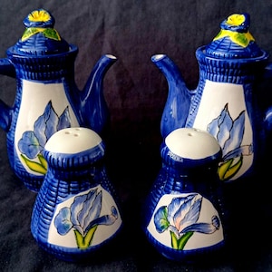 Op de afbeelding: Een set van twee keramische theepotten en twee bijpassende zout- en pepervaatjes, allemaal voorzien van een diepblauw en wit kleurenschema met handgeschilderde blauwe irismotieven en gele accenten op de deksels, geplaatst op een donkere ondergrond.