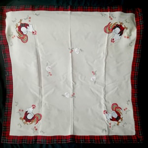 Puede incluir: Un paño de tela cuadrado de color blanco roto con un borde de cuadros rojos y negros. El centro está decorado con cuatro gallos bordados en las esquinas, cada uno con plumas de cola rojas y verdes, junto a pequeños motivos de huevos y flores.
