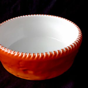 Könnte beinhalten: Eine runde Keramikschale mit glatter weißer Innenseite und leuchtend orangefarbener Außenseite. Der Rand weist eine ausgeprägte, erhabene seilartige Textur mit abwechselnden weißen und orangefarbenen vertikalen Streifen auf.