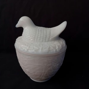 Op de afbeelding: Een witte, vintage-stijl dekselschaal in de vorm van een vogel op een nest. De vogel heeft gedetailleerde veren en een licht geopende snavel. De schaal heeft een textuur, geweven mandontwerp.