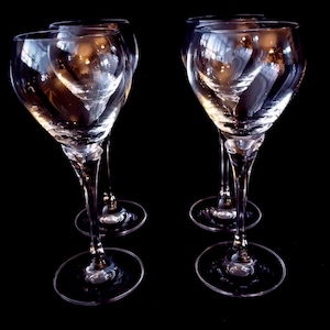 Op de afbeelding: Vier heldere wijnglazen met lange stelen en ronde voetjes tegen een zwarte achtergrond. De glazen hebben een klassiek ontwerp met een afgeronde kelk en een slanke steel, die licht reflecteert.