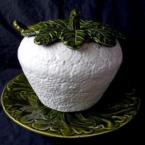 May include: A white ceramic decorative jar with a textured surface, topped with a green leaf-shaped lid. The jar sits on a matching green leaf-patterned plate. The overall design is reminiscent of a stylized fruit.