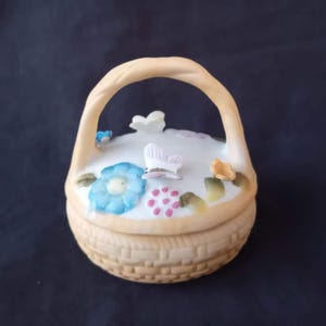Op de afbeelding: Een kleine keramische mand met een handvat, versierd met een lichtblauwe bloem, roze bessen en vlinders. De mand heeft een geweven textuur en een crèmekleurig handvat en basis. De deksel is wit met bloemaccenten.