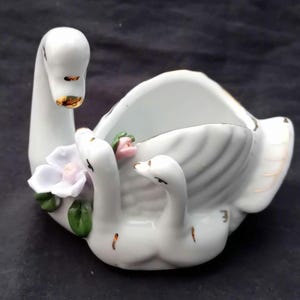 Op de afbeelding: Een witte keramische zwaanfiguur met drie jongen, een bloem en gouden accenten. De zwaan heeft zijn kop omhoog en de jongen zijn eronder genesteld. De figuur staat op een donkere achtergrond.