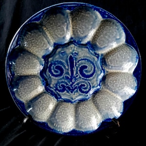 Op de afbeelding: Blauw en wit keramisch oesterbord met een centraal fleur-de-lis-ontwerp. Het bord heeft twaalf afzonderlijke oestercompartimenten en een glanzende afwerking. Het bord is rond en heeft een opstaande rand.
