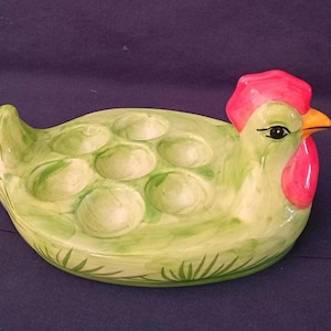 Op de afbeelding: Een keramische eierhouder in de vorm van een kip. De eierhouder is groen geschilderd met een rode kam en lellen, en een gele snavel. De houder heeft zeven ronde uitsparingen voor eieren.