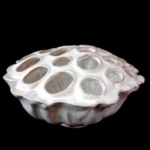 Op de afbeelding: Een witte decoratieve keramische kom met een uniek, poreus ontwerp. De kom heeft talrijke ovale openingen over het oppervlak en een golvende, getextureerde rand. De kom staat tegen een zwarte achtergrond.