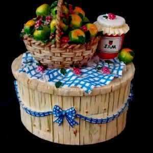 Op de afbeelding: Een decoratieve houten doos met een fruitmand en een pot jam. De mand is gevuld met groene en oranje vruchten en rode bessen. De jam pot heeft een rood etiket met het woord "JAM". Een blauw-wit geruit kleed en een blauw lint versieren de doos.