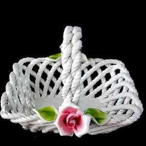Op de afbeelding: Een witte keramische mand met een geweven ontwerp en een gevlochten handvat. De mand is versierd met een roze roos en groene bladeren. De mand wordt waarschijnlijk gebruikt voor decoratieve doeleinden.