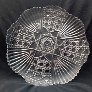 Op de afbeelding: Transparante glazen serveerschaal met een ster- en geometrisch patroon. De schaal heeft een geschulpte rand en een gedetailleerd ontwerp van stralende lijnen en cirkelvormige vormen. Een decoratief stuk om te serveren of tentoon te stellen.