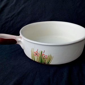 Op de afbeelding: Witte keramische kom met een handvat, met een bloemenontwerp van roze bloemen en groen gras. Het handvat is donkerbruin. De kom is geschikt om te serveren of te koken.