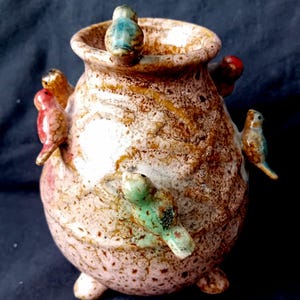 Pode incluir: Um vaso de cerâmica redondo com um esmalte salpicado em rosa e castanho, apresentando três pequenos pés na base. Vários pássaros pequenos esculpidos em várias cores, incluindo vermelho, verde e azul, estão fixados na superfície exterior e no rebordo.