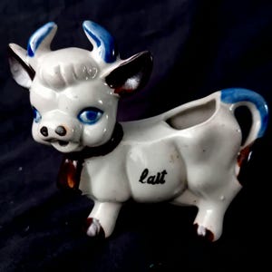 Op de afbeelding: Een keramische koeienroomkannetje met een witte body, blauwe hoorns en zwarte en bruine accenten. Het woord "lait" staat op de zijkant. Het roomkannetje heeft een handvat en een schenktuit.