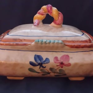 Op de afbeelding: Een keramische sieradendoos met deksel, met een bloemenontwerp met blauwe en roze bloemen. De doos heeft een crèmekleurige basis met een decoratieve handgreep in geel, roze en oranje tinten. De doos is rechthoekig met afgeronde randen.