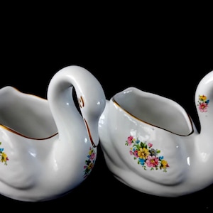 Op de afbeelding: Twee witte keramische zwanenvormige plantenbakken met gouden rand. Elke plantenbak heeft een bloemenontwerp in roze, geel, blauw en groen. De zwanen hebben gebogen halzen en open bakken.