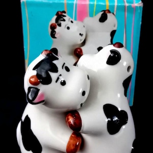 May include: A set of three ceramic cow figurines, each with a white body, black spots, and brown accents. The cows are hugging each other. The figurines are in front of a blue box with pink and white stripes.