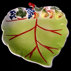 Op de afbeelding: Een keramische serveerschaal in de vorm van een blad, met een getextureerd groen oppervlak en rode aderdetails. De rand is wit. De schaal is versierd met blauwe druiven, gele peren en groene bladeren. Een decoratief stuk.