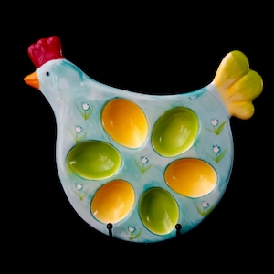 Op de afbeelding: Een keramische eierhouder in de vorm van een kip, lichtblauw geschilderd met gele en groene eivormige compartimenten. De kip heeft een rode kam, een oranje snavel en gele staartveren. Kleine witte bloemaccenten.
