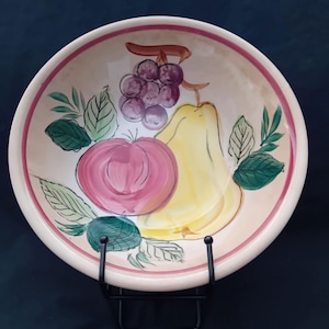 Può includere: Una ciotola da portata in ceramica dipinta a mano con un disegno di frutta. La ciotola presenta un bordo rosa e un interno beige con illustrazioni di una pera gialla, uva viola e una mela rosa. Le foglie verdi accentuano la frutta.