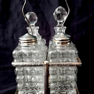 Op de afbeelding: Een kruidenset bestaande uit vier vierkante, heldere glazen flessen met een gestructureerd ruitpatroon, geplaatst in een zilverkleurige metalen houder met een gebogen handvat. Twee flessen hebben gefacetteerde glazen stoppen en twee metalen deksels.