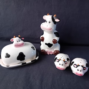 Può includere: Un set da cucina a tema mucca in ceramica. Il set comprende una burriera, una statuina di mucca seduta e due saliere e pepiere. Gli articoli sono bianchi con macchie nere e dettagli marroni. La burriera è di forma ovale.