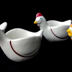 Puede incluir: Un juego de cuatro hueveras de cerámica blanca con forma de gallina. Cada huevera presenta una línea roja pintada a mano en el lateral, con detalles faciales distintivos que incluyen crestas rojas y picos de color amarillo o rojo.