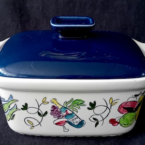 May include: A rectangular white ceramic casserole dish with a deep blue lid and two side handles. The exterior features a colorful hand-painted design of a wine bottle, grapes, a wine glass, cherries, and green leaves against a white background.