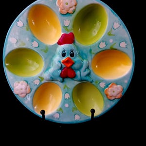 May include: A ceramic egg serving platter with a light blue background, featuring a cartoon chicken in the centre and egg-shaped compartments in yellow and green. The platter is decorated with white flowers and tulips.