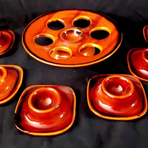 May include: A set of vintage ceramic egg cups and a serving platter. The egg cups are square with a rounded top, and the platter has multiple circular indentations. The set is glazed in shades of brown and orange.