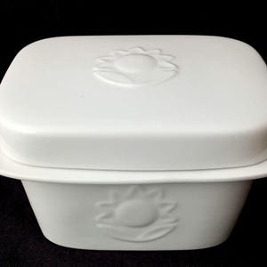 Op de afbeelding: Een witte keramische botervloot met een rechthoekige basis en een bijpassend deksel. Zowel het deksel als de voorzijde van de basis zijn voorzien van een verhoogd, in reliëf aangebracht zonnebloemmotief. Het object heeft gladde, ronde randen.