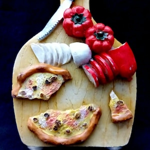 May include: A decorative wooden cutting board with a knife, red bell peppers, sliced onions, mozzarella, and pizza slices. The board has two metal hooks at the bottom. The image is set against a dark background.