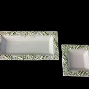 Op de afbeelding: Twee witte keramische serveerschalen. Eén is rechthoekig, de andere vierkant. Beide hebben een verhoogd druiven- en bladerontwerp langs de randen in een gedempte groene kleur. De schalen staan tegen een zwarte achtergrond.