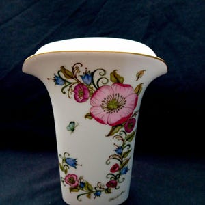 Op de afbeelding: Een witte keramische vaas met een uitlopende bovenkant, versierd met een bloemenontwerp. De bloemen zijn roze, blauw en rood, met groene bladeren en een vlinder. De rand is afgewerkt met goud.