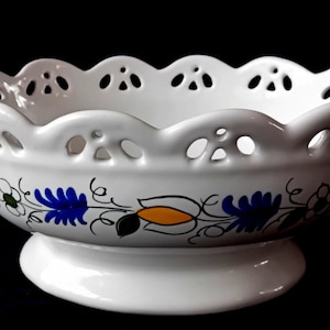 Op de afbeelding: Witte keramische kom met een geschulpte rand en decoratief uitgesneden ontwerp. De kom heeft een bloemenpatroon in blauw, geel en groen. Hij staat op een ronde voet.
