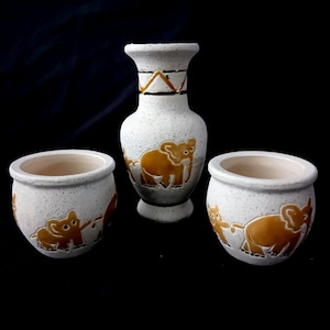 Op de afbeelding: Een set van drie keramische vazen en potten. De stukken zijn wit met een gespikkelde textuur en hebben een herhalend ontwerp van bruine olifanten. De hoogste vaas heeft een smalle hals met geometrische accenten.