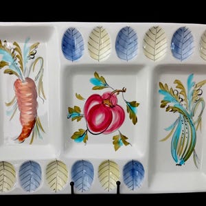 May include: A rectangular white ceramic serving tray with three divided sections. The left section features a painted orange carrot, the center a red fruit, and the right a green vegetable. Twelve small oval indentations with blue and yellow leaf patterns line the top and bottom edges.