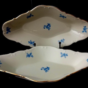 Op de afbeelding: Twee ovale serveerschalen van wit porselein met geschulpte randen en goudkleurige afwerking. Elke schaal is voorzien van een delicaat patroon van kleine blauwe bloemclusters verspreid over het binnenoppervlak, wat zorgt voor een strakke uitstraling.