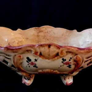 Puede incluir: Un cuenco de cerámica ovalado y antiguo con borde festoneado. El cuenco es de color crema con ribete rojo y marrón. Presenta diseños florales y descansa sobre tres pequeños pies. El diseño del cuenco sugiere que es vintage.