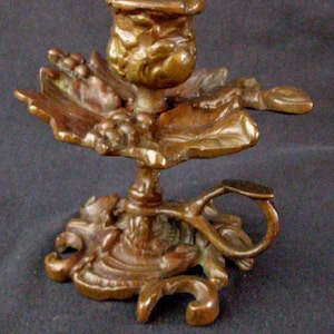 Op de afbeelding: Een sierlijke bronzen kandelaar met een gedetailleerd bloemen- en bladerontwerp. De kaarsenhouder is omgeven door gebeeldhouwde bladeren en de basis heeft ingewikkeld krulwerk. Een klein handvat is aan de zijkant bevestigd.