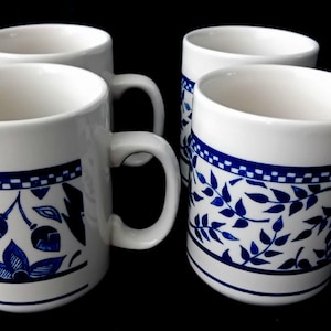 Op de afbeelding: Een set van vier witte keramische mokken met een blauw patroon. Elke mok heeft een handvat en een decoratieve band bij de bovenrand met een geblokt ontwerp. De onderste delen tonen blauwe bloem- en bladmotieven en twee dunne blauwe lijnen.