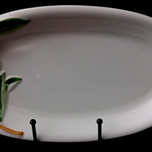 Może przedstawiać: Owalny biały półmisek ceramiczny z wypukłym, trójwymiarowym wzorem po lewej stronie. Relief przedstawia gałązkę z dwiema ciemnofioletowymi oliwkami, jedną jasnozieloną oliwką oraz kilkoma zielonymi liśćmi z ciemnymi krawędziami.