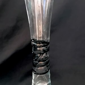 Op de afbeelding: Een heldere glazen vaas met een uitlopende bovenkant en een zwart, wervelend ontwerp rond het midden. De vaas heeft een slanke, elegante vorm en een heldere basis. Het zwarte ontwerp voegt een decoratief element toe.