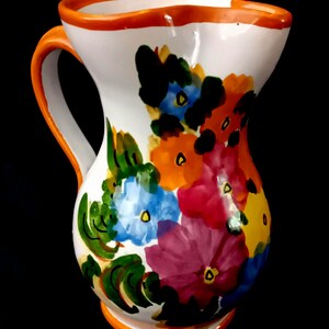 Op de afbeelding: Witte keramische kan met een levendig bloemmotief. De kan heeft een feloranje handvat en rand, met kleurrijke bloemen in tinten blauw, rood, oranje en paars. De kan is een decoratief item.