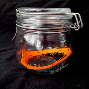 Op de afbeelding: Een doorzichtige glazen voorraadpot met een metalen beugel en een hefboomsluiting. De pot heeft een feloranje geschilderde band rond het midden met de in reliëf aangebrachte tekst "Le Parfait Super" zichtbaar op het glazen oppervlak.