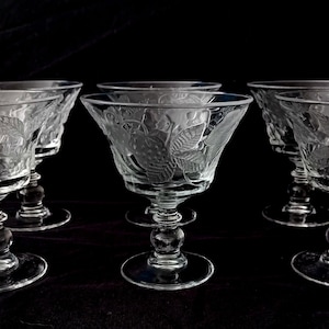 Op de afbeelding: Set van zes heldere, geëtste champagnecoupeglazen. Elk glas heeft een brede kom met een blad- en bessenontwerp, ondersteund door een steel met een decoratieve bal en een cirkelvormige voet. De glazen staan tegen een donkere achtergrond.