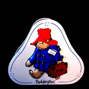 May include: A white, triangular tin with a cartoon illustration of Paddington Bear. The bear is wearing a blue coat and red hat, carrying a suitcase. The tin is labelled with the word "Paddington" at the bottom.