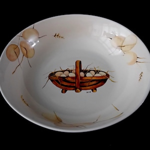 May include: A white ceramic serving bowl with a hand-painted design. The bowl features a brown basket filled with white eggs, surrounded by painted foliage. The rim of the bowl has additional painted floral accents. The bowl is set against a black background.