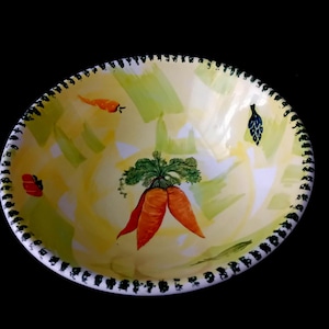 Op de afbeelding: Een handbeschilderde keramische kom met een gele binnenkant en een zwart-witte rand. De kom heeft een grillig ontwerp van wortelen, bladeren en andere groente-thema accenten. Perfect voor het serveren van salades of als decoratief stuk.
